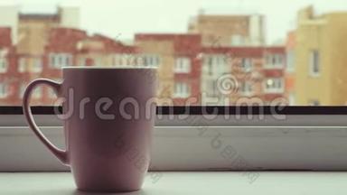 在白雪皑皑的冬天早晨，热气腾腾的一杯茶站在窗户上。 舒适的粉红咖啡杯。 大雪纷飞，大雪纷飞